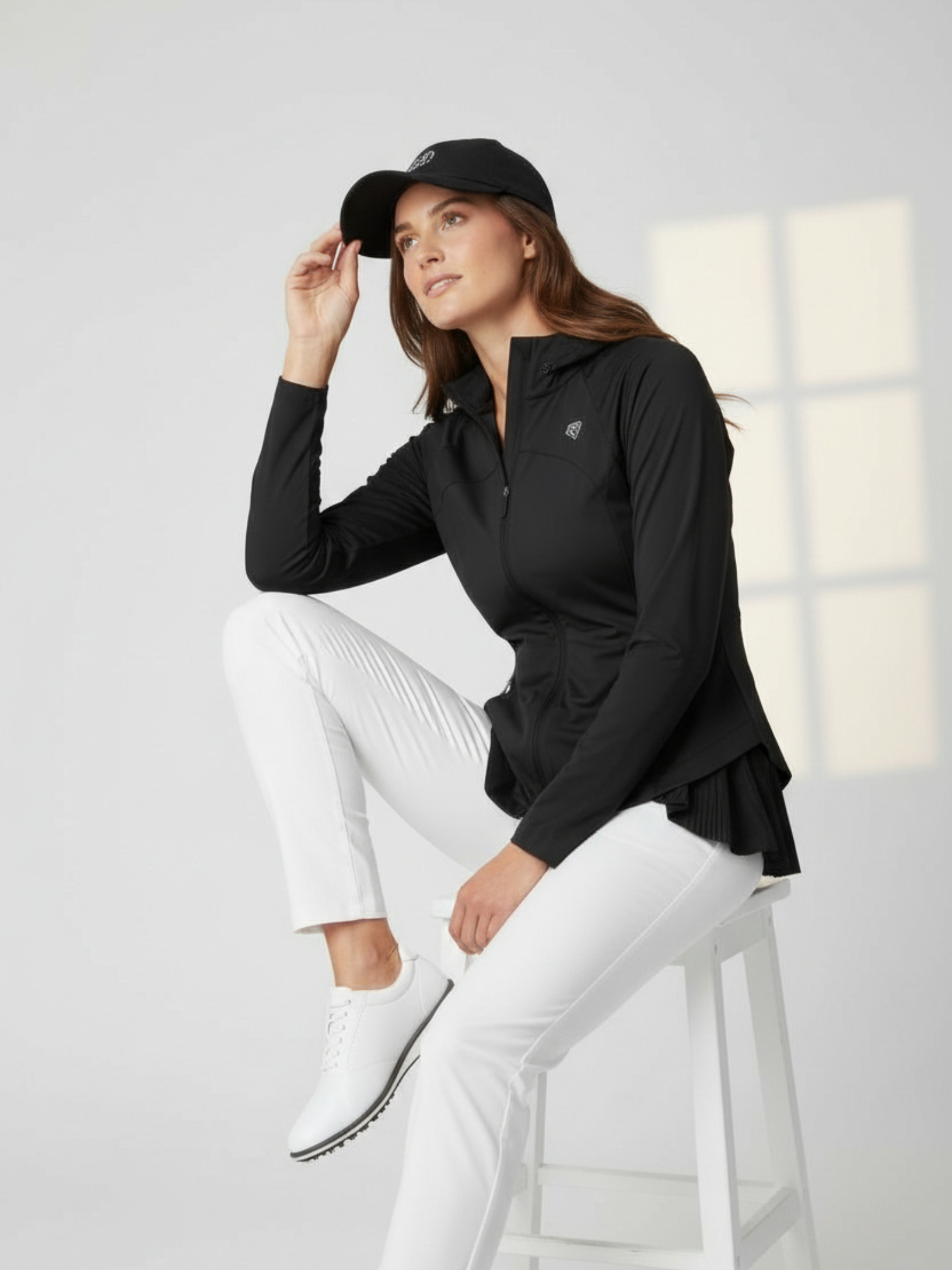 Woman wearing a black NGA Backtalk athletic jacket and white pants sitting on a stool against a light gray background