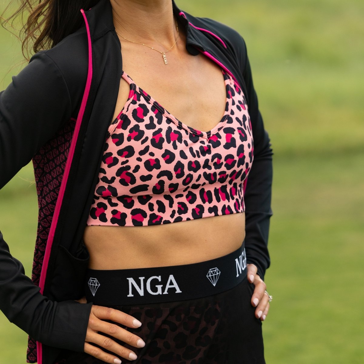A woman wearing a pink and black leopard-print NGA Ta-Tas bra and matching Sheer Courage mini skirt with a black waistband featuring 'NGA' branding, posed outdoors on a golf course.